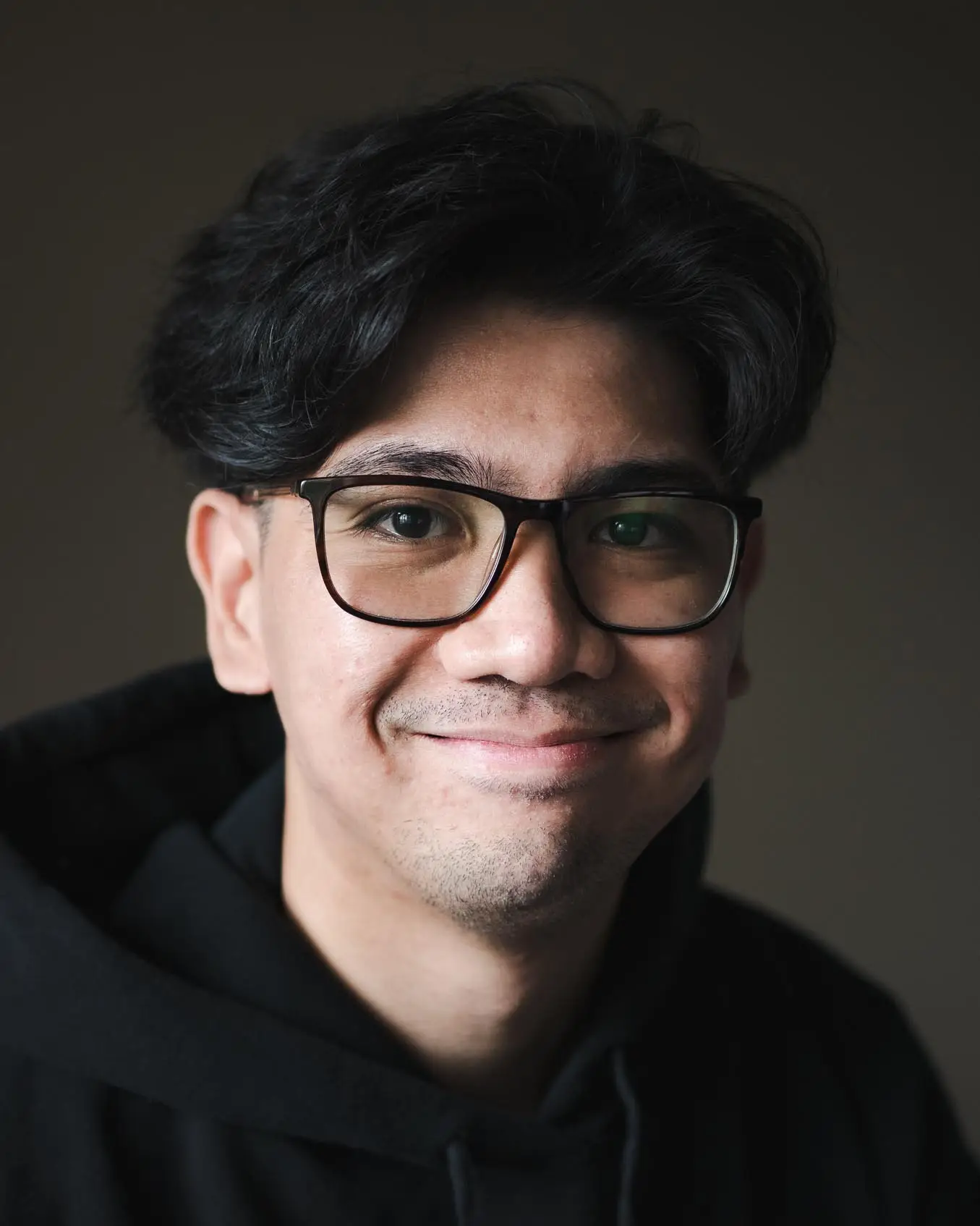 Alec Hernaez smiling in a black hoodie and black framed glasses